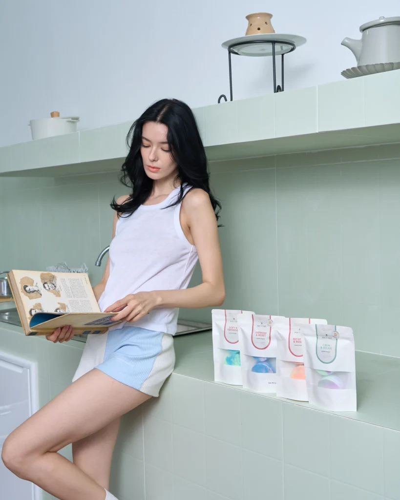 Woman reading in a green-tiled bathroom beside Holy Fizz shower steamers in Enegize & Breathe Soft & Feminine, Refresh & Reset, and Calm & Relax scents on a green tiled ledge.
