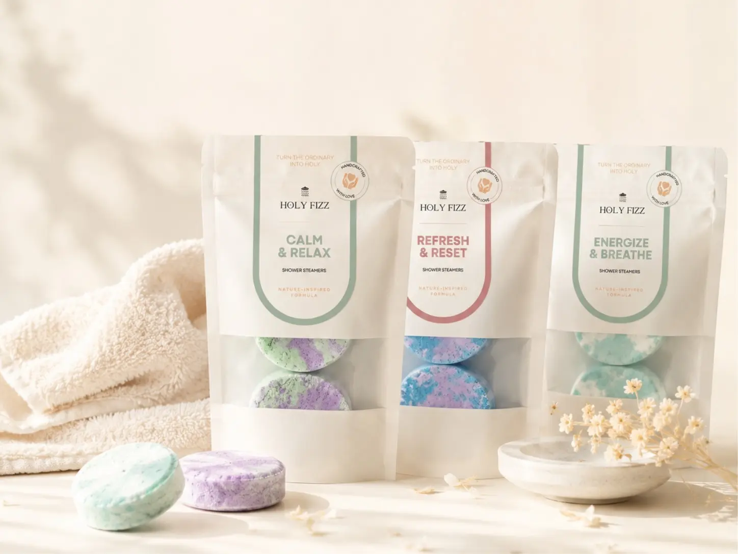 ซองผลิตภัณฑ์ Holy Fizz Shower Steamer 3 กลิ่น Calm & Relax, Refresh & Reset และ Energize & Breathe วางคู่กับผ้าขนหนูและก้อนอโรม่าสีเขียวมิ้นต์และม่วง