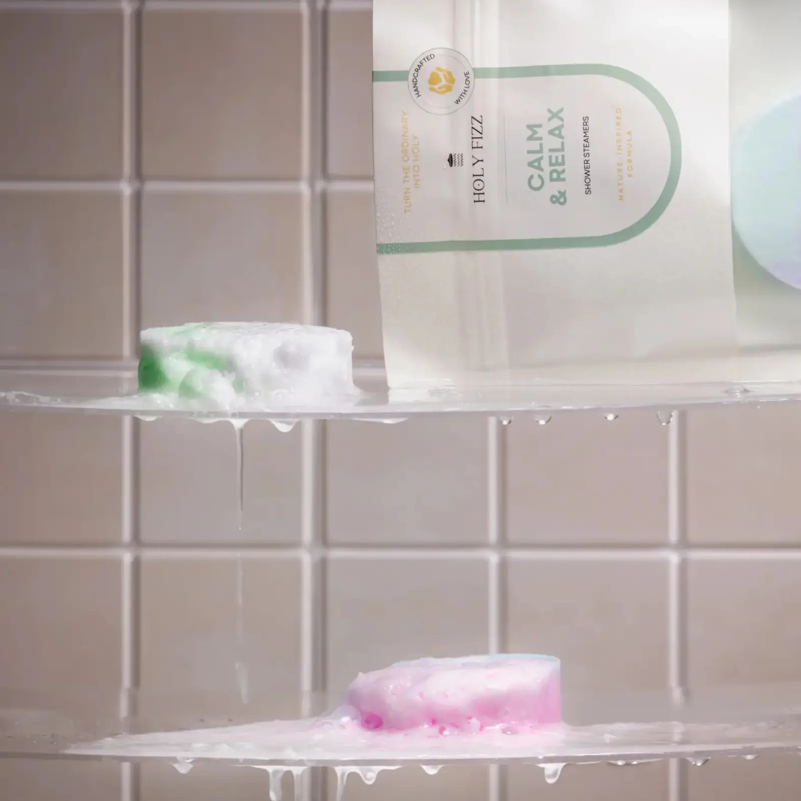 ชาวเวอร์สตีมเมอร์ Holy Fizz Calm & Relax บนชั้นวางในห้องอาบน้ำ พร้อมชาวเวอร์สตีมเมอร์สีเขียวและชมพูที่กำลังละลายน้ำ