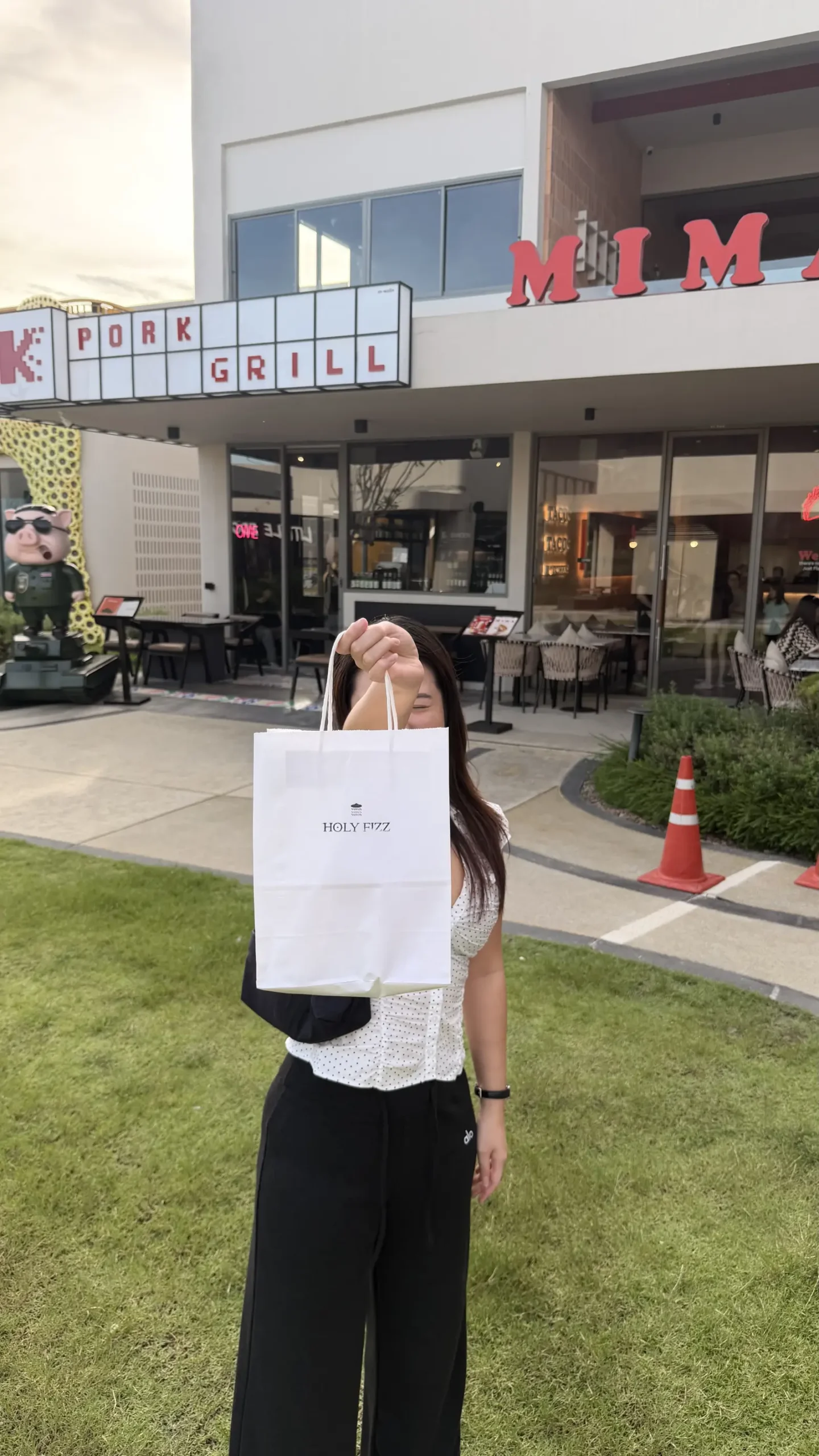 Woman holding a Holy Fizz Thailand shopping bag in front of a restaurant and community mall.