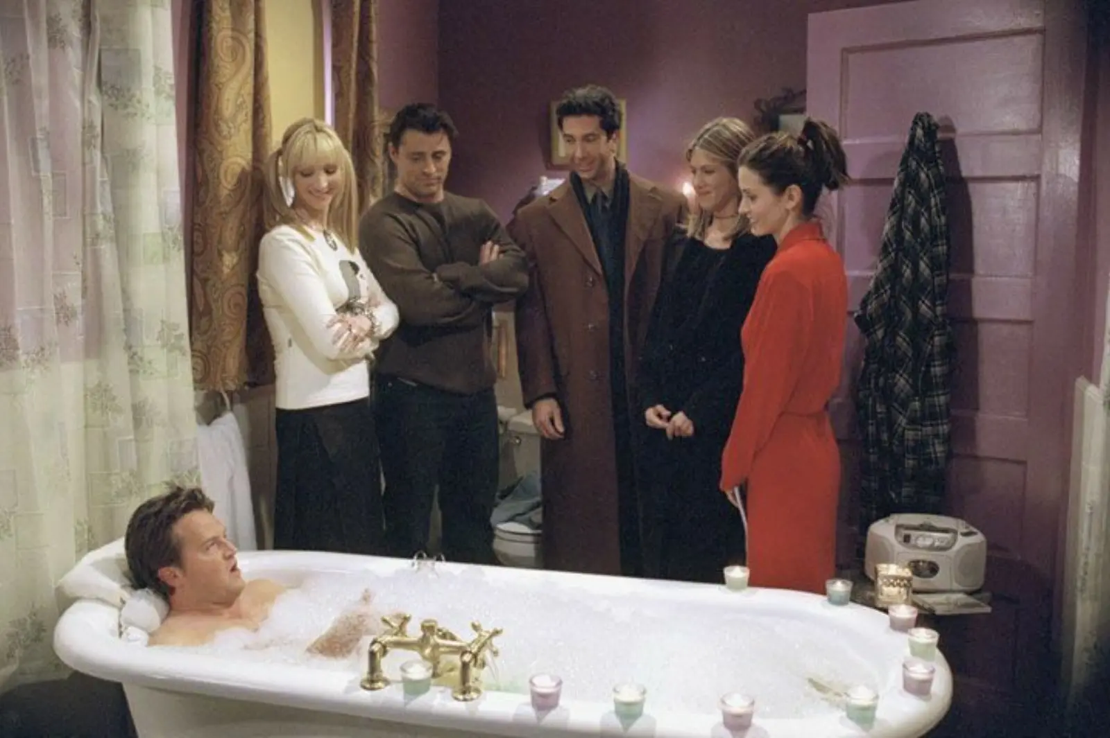 Group of friends gathered around a man relaxing in a bubble bath with candles in a bathroom