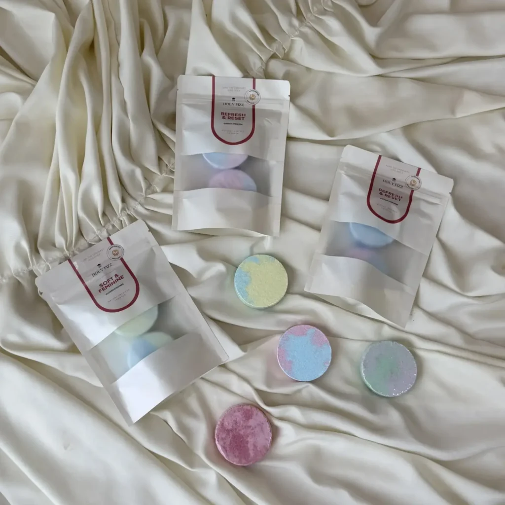Holy Fizz Shower Steamers in pastel colors with Refresh & Reset and Soft & Feminine pouches on satin fabric
