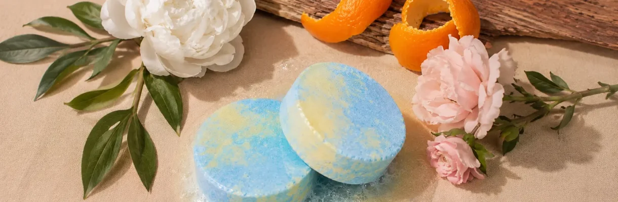Holy Fizz Peony Musk blue and yellow shower steamers fizzing beside white peony, pink roses, orange peel and green leaves on linen